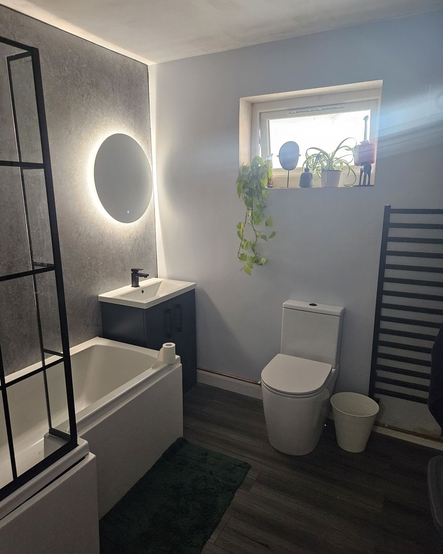 A finished bathroom with a black-framed bath screen, a round lit mirror, a navy basin unit, a toilet and a black towel radiator