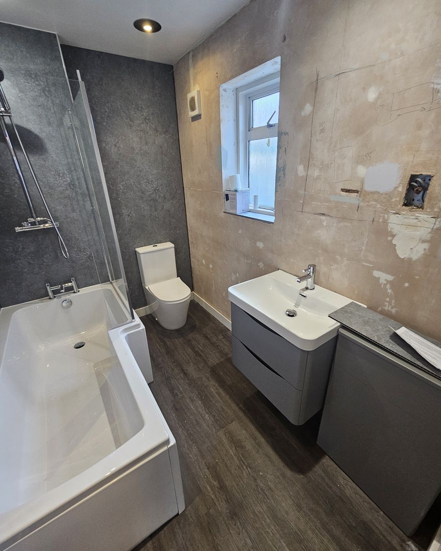 A shower-bath with a glass screen, a grey basin unit and a toilet, with the walls in bare plaster ready for decorating