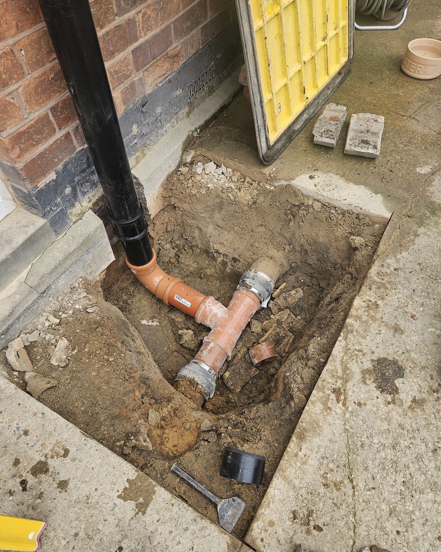A new black soil pipe running down the back wall into new orange drain fittings in a dug-out section of the yard