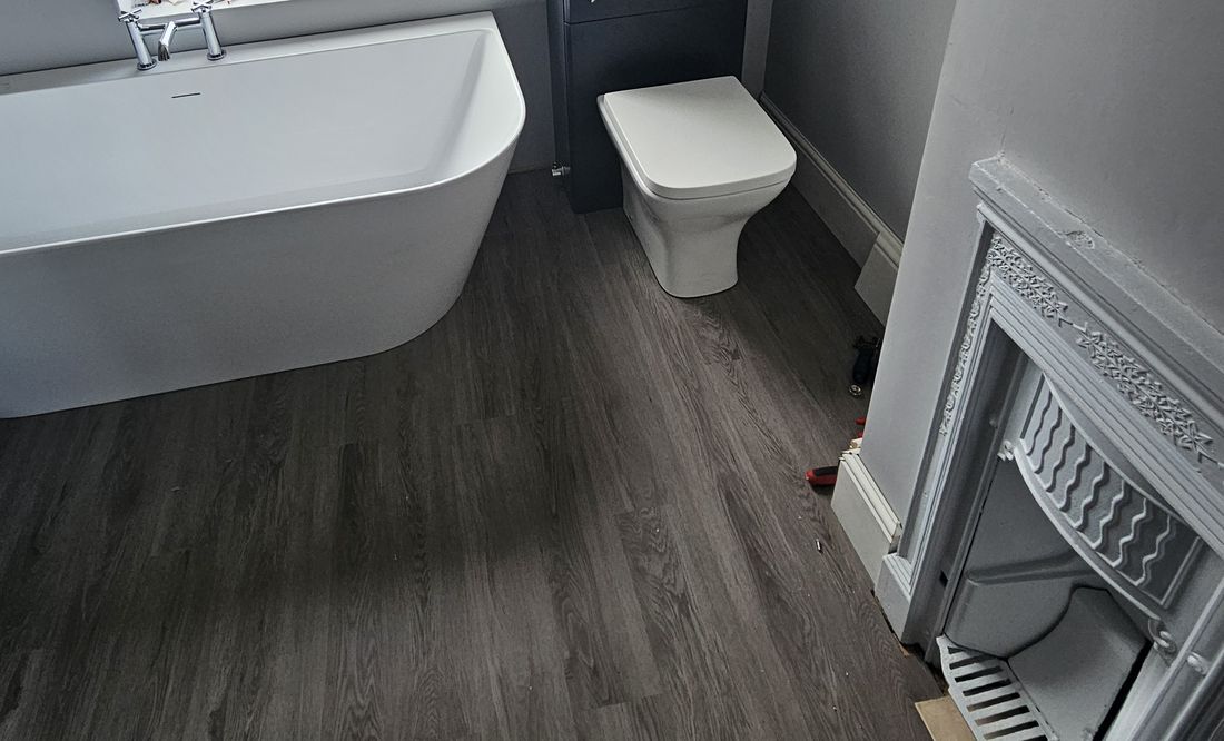 The finished bathroom: a freestanding bath, a toilet with a navy cistern unit, a grey plank floor and a cast-iron fireplace