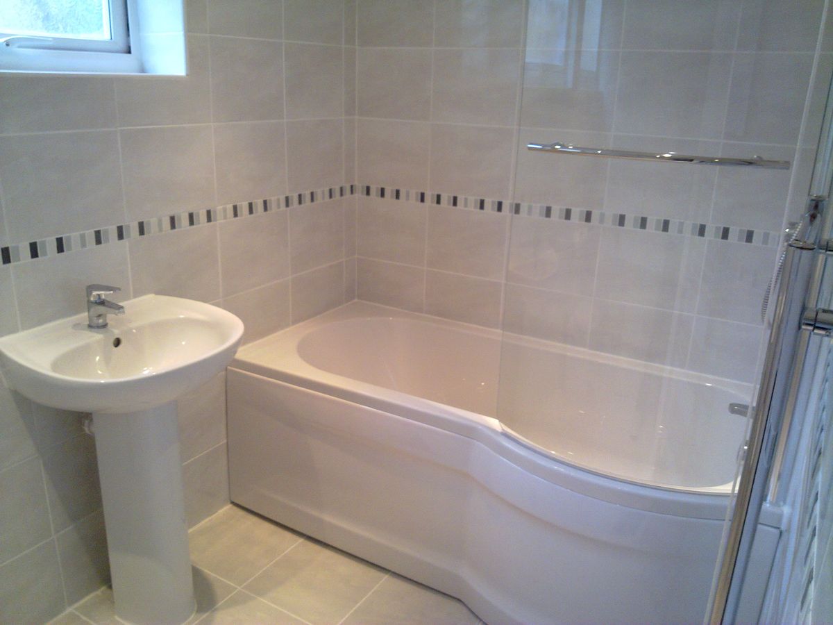 A tiled bathroom with a P-shaped bath, a pedestal basin and a decorative tile border