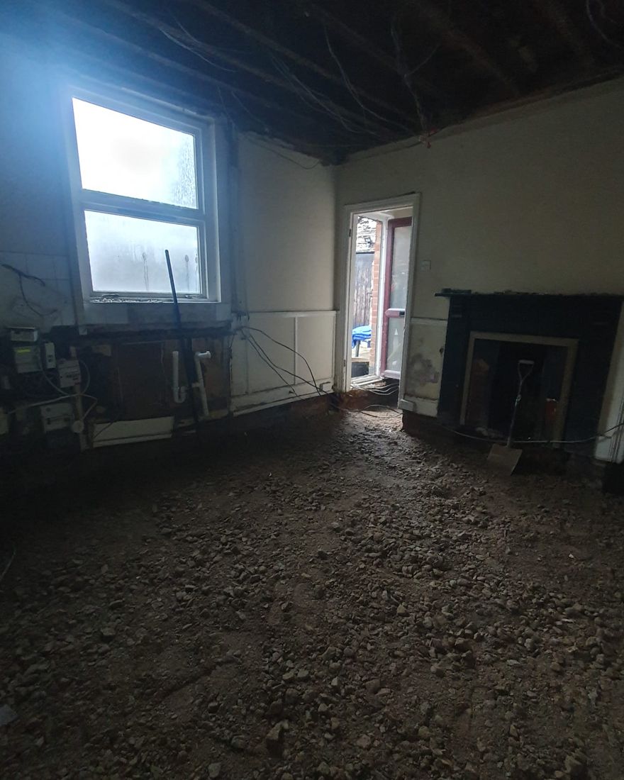 Kitchen-diner with the ceiling taken down to the timber joists and the floor dug out, a window on the left and a fireplace on the right