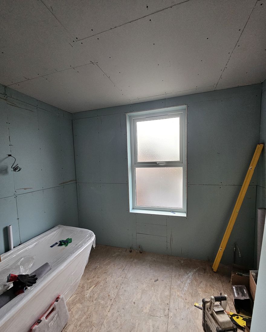 Bathroom ceiling boarded in plasterboard above an obscured-glass window, walls boarded in green moisture-resistant board, bath under protection