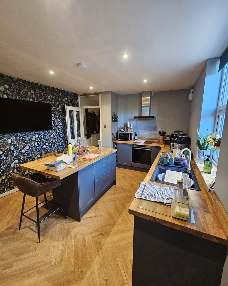 The finished kitchen-diner: dark blue island, oak worktops, a herringbone floor and a wallpapered feature wall