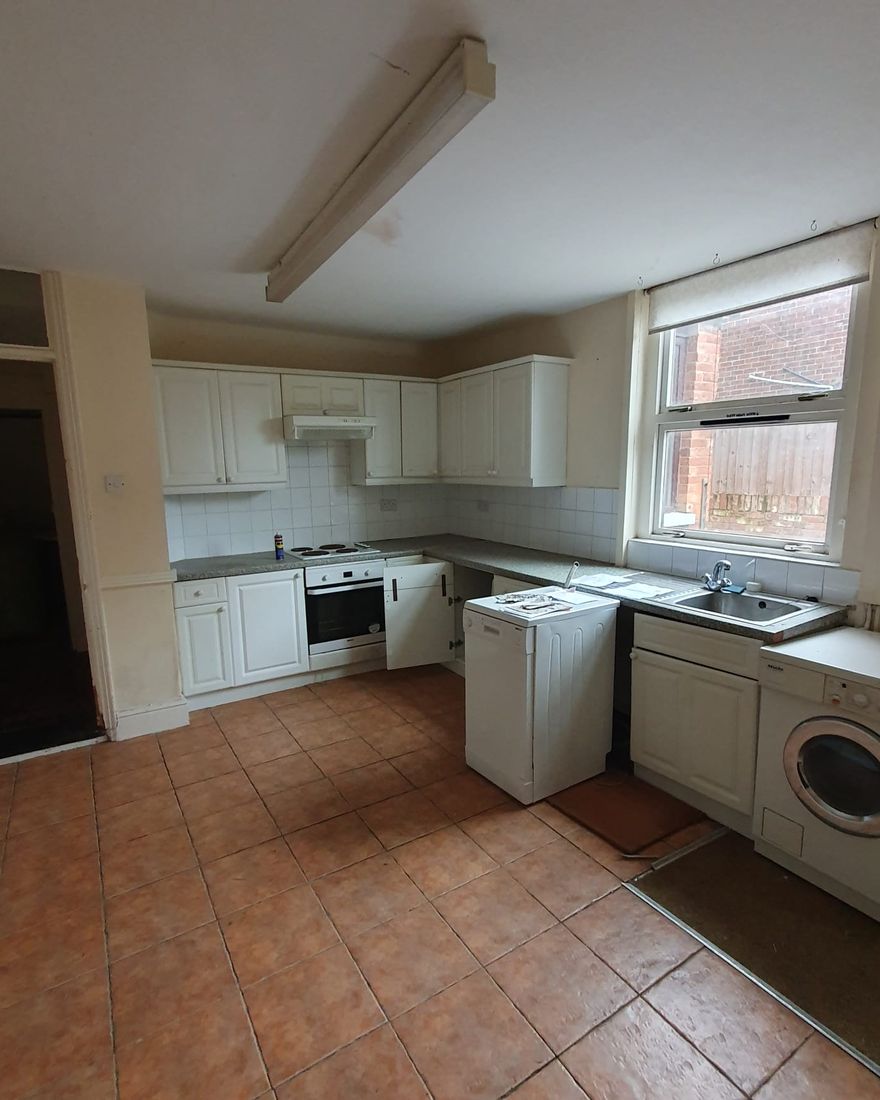The old kitchen: white units, terracotta floor tiles and a washing machine under a fluorescent strip light
