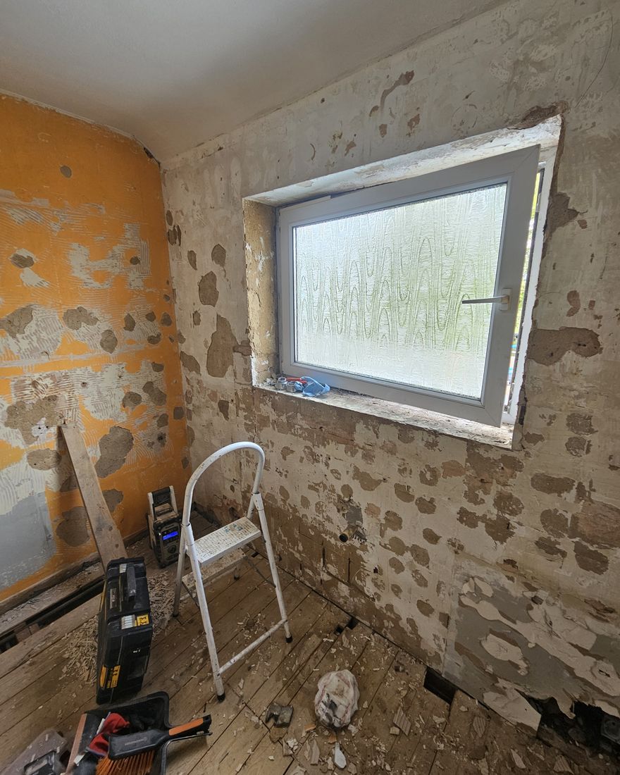 Small room stripped to old plaster and orange tile adhesive under an obscured-glass window
