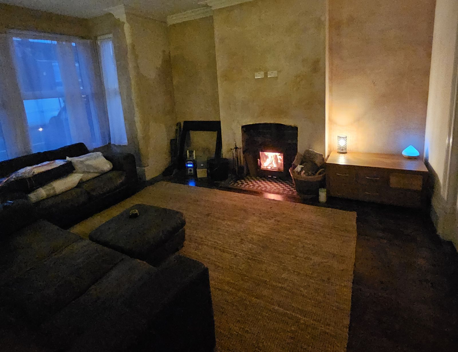 Front room in the evening, with a log burner lit in the re-plastered chimney breast and the bay window to the left