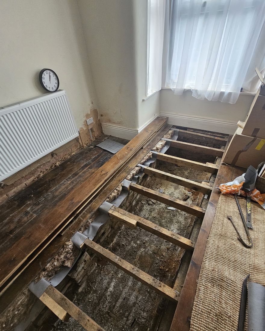 Front-room floorboards lifted, showing the joists, beside the radiator and the bay window